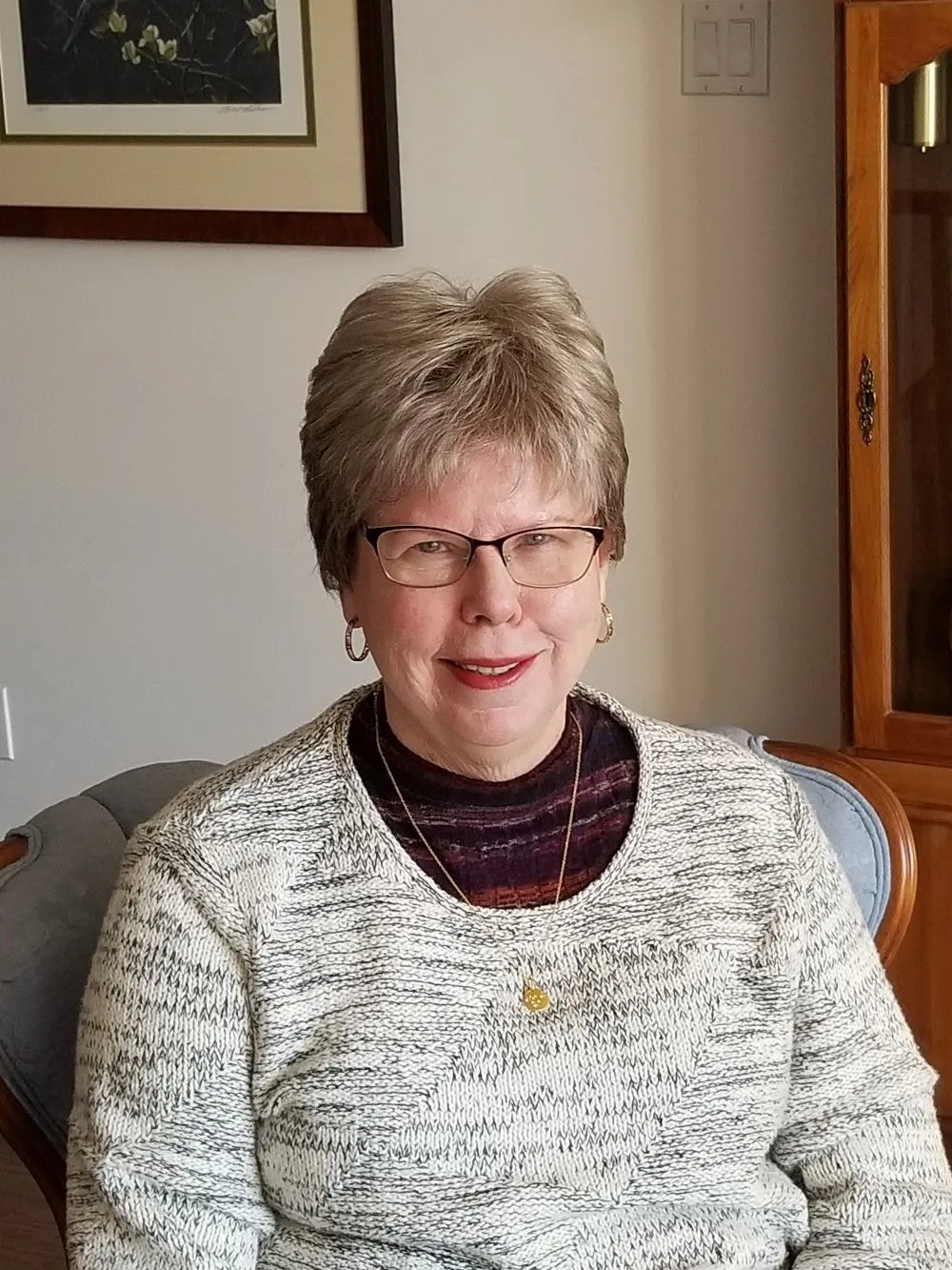 Heather siting on a sofa in her living room. She has short white hair, glasses and wears a grey jumper over a black top